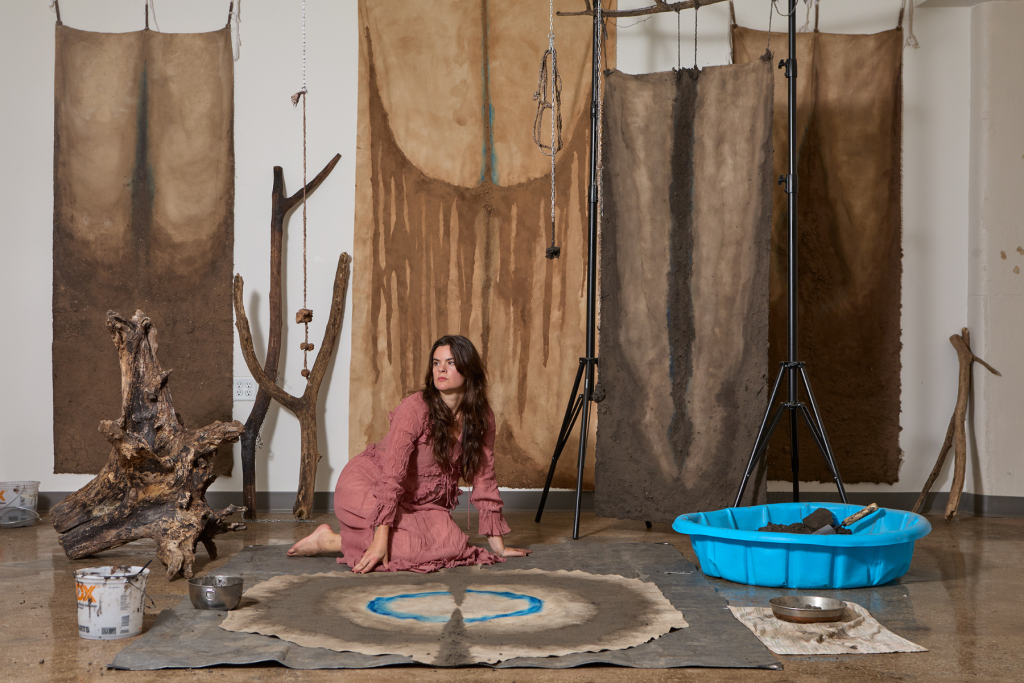 Artist Larissa Belcic kneels amid an installation of earth-toned hanging textiles, branches, roots, chains, and soil, with a circular blue-marked work on the floor in front of her.