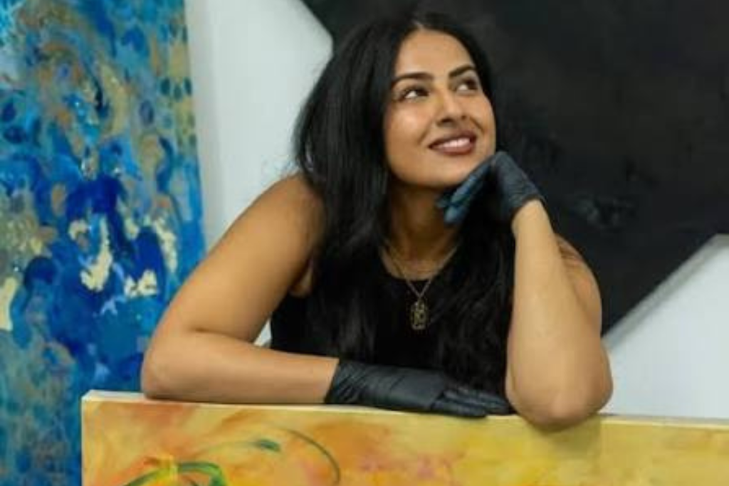 Smiling artist with long dark hair and black gloves rests her arms on a warm-toned painting, surrounded by large abstract works in vivid blue, gold, black, and white