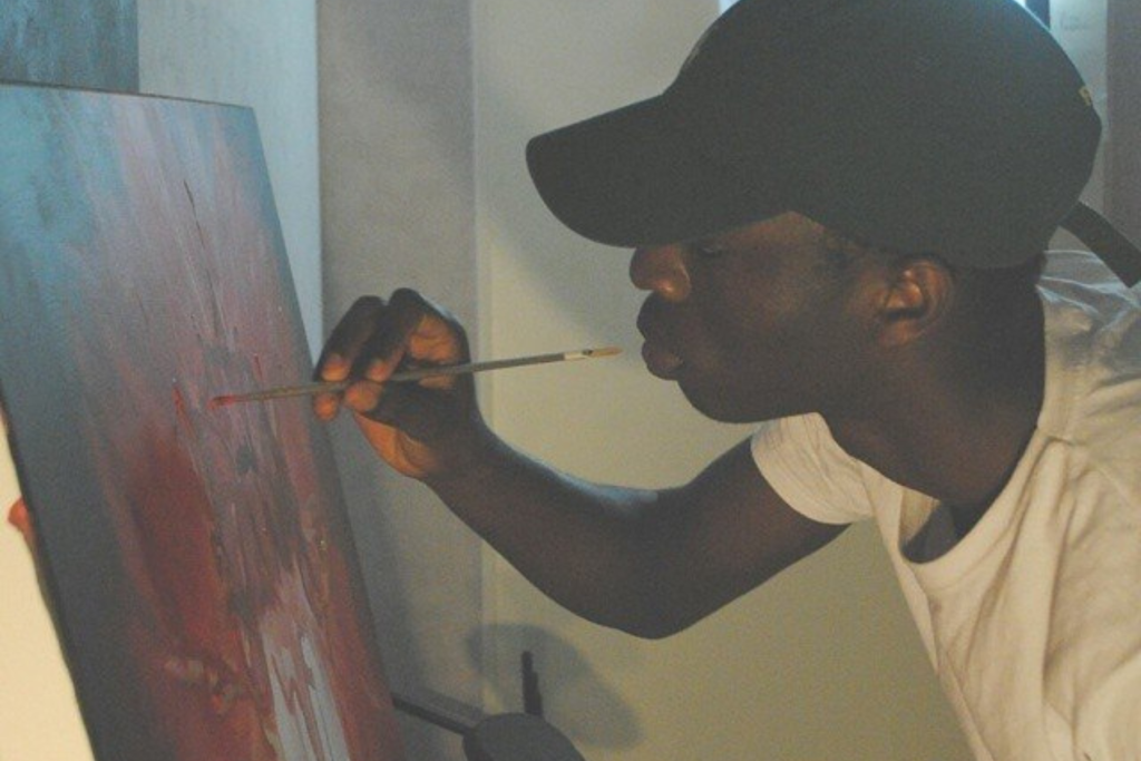 ARTS 14C Special Project artist Lossapardo painting on canvas, wearing a black baseball cap and white T-shirt while working closely with a fine brush.