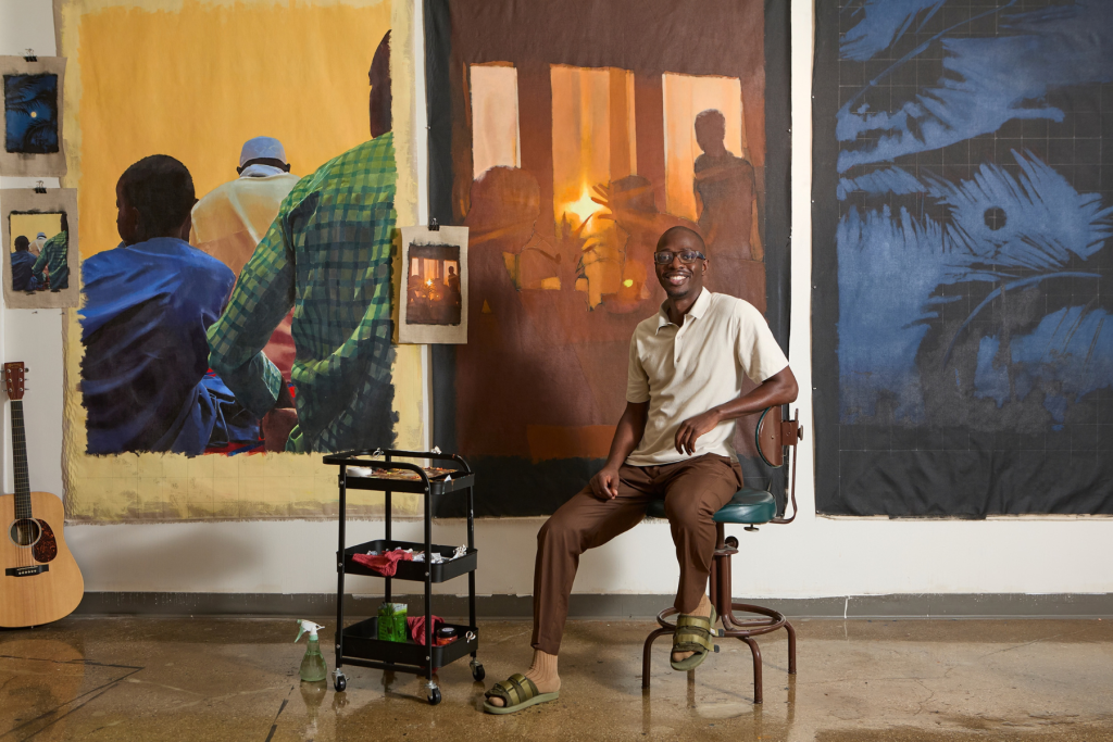 Artist Lossapardo sits smiling in his ARTS 14C studio, surrounded by large-scale figurative paintings in rich blues, browns, greens, and golds, with a guitar and painting supplies nearby.