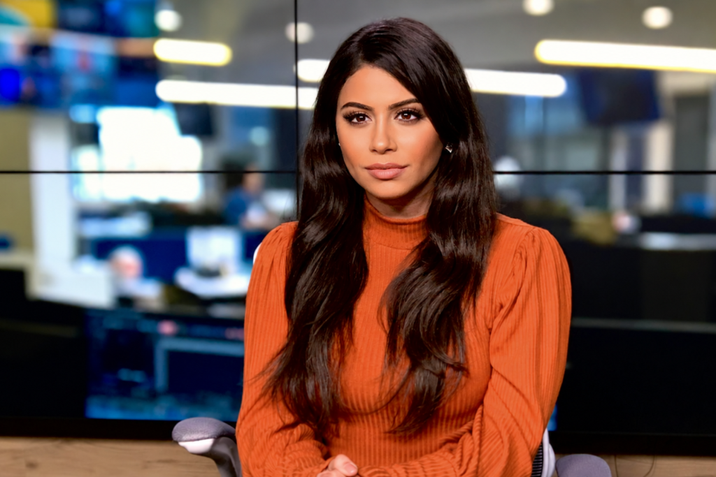 Woman in an orange sweater seated in a newsroom studio, looking toward the camera with softly blurred monitors and lights in the background.