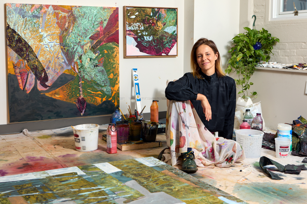 Artist Luiza Gottschalk sits among paints, brushes, and works in progress in her ARTS 14C studio, surrounded by richly layered abstract paintings and colorful materials.