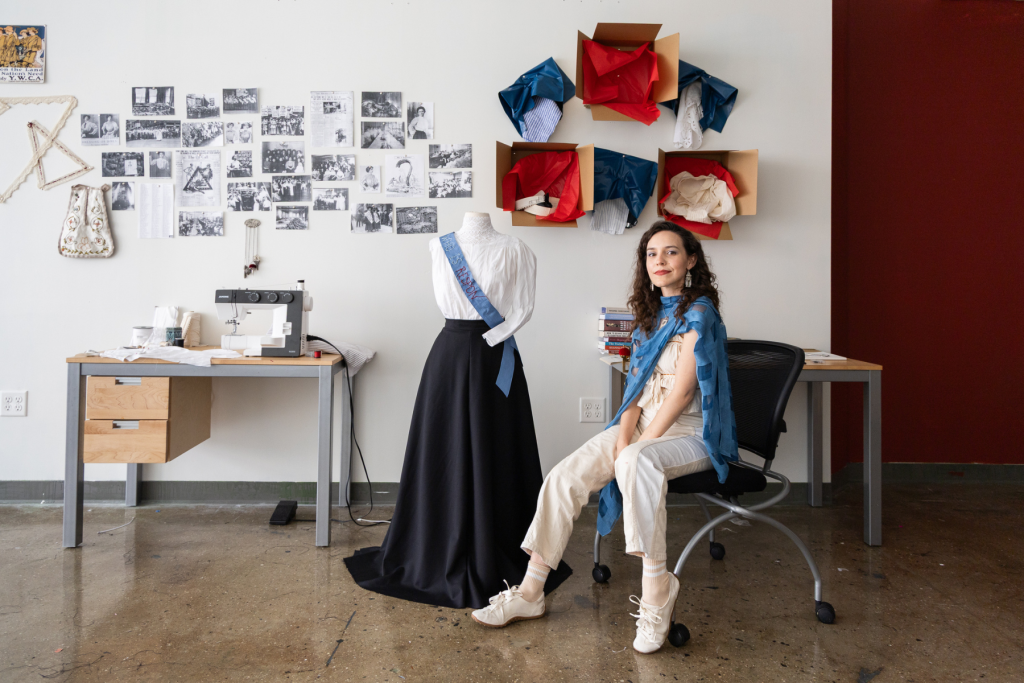 Artist Madison Bouse sits in her studio beside a dress form wearing a white blouse, long black skirt, and blue sash, surrounded by sewing materials, archival photographs, and textile-based installations.