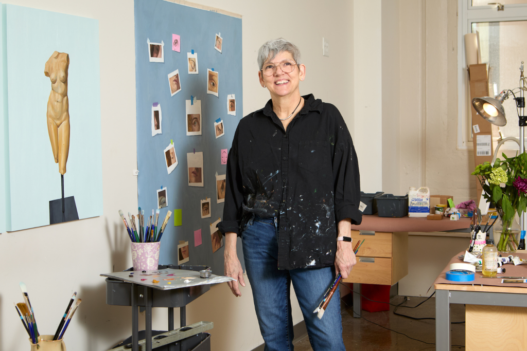 Artist Margaret Murphy stands holding paintbrushes in her ARTS 14C live/work studio, surrounded by painting supplies and works in progress, including studies of the human figure and closely observed facial features.
