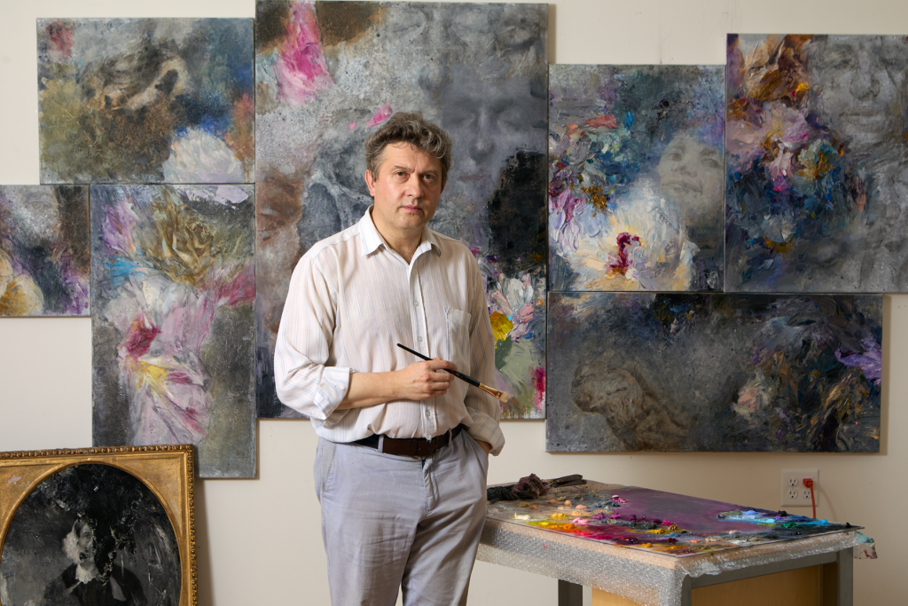 Fellowship Artist Mark Shilenkov stands in his ARTS 14C studio holding a paintbrush, surrounded by layered abstract and figurative paintings in muted grays with vivid touches of pink, blue, and yellow.