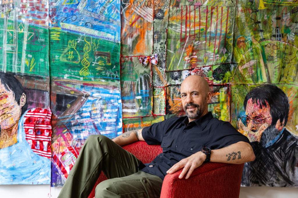 Artist Mateo Gutierrez sits in a red chair in front of large, densely layered mixed-media works filled with vivid color, gestural marks, collage, and expressive figures.