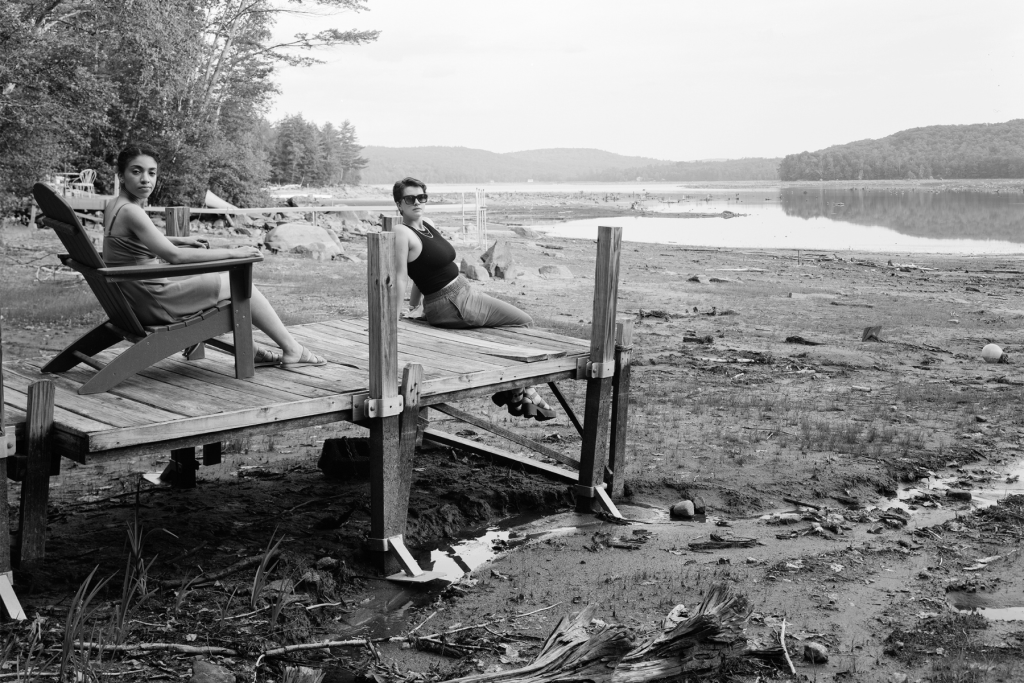 Black-and-white photograph by Matthew Cohn of two people relaxing on a wooden dock beside a low-water lake, with a wooded shoreline and hills stretching across the distance.