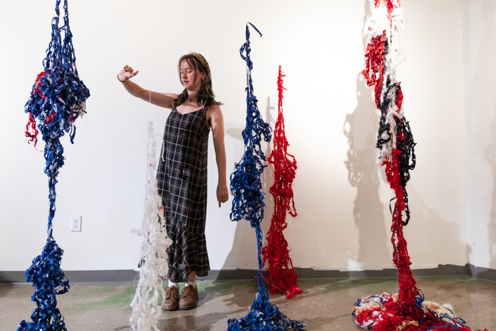 Artist Maud Etheridge stands among suspended fiber sculptures made of loosely woven red, blue, white, and black strips, their elongated forms casting shadows across the ARTS 14C studio wall.