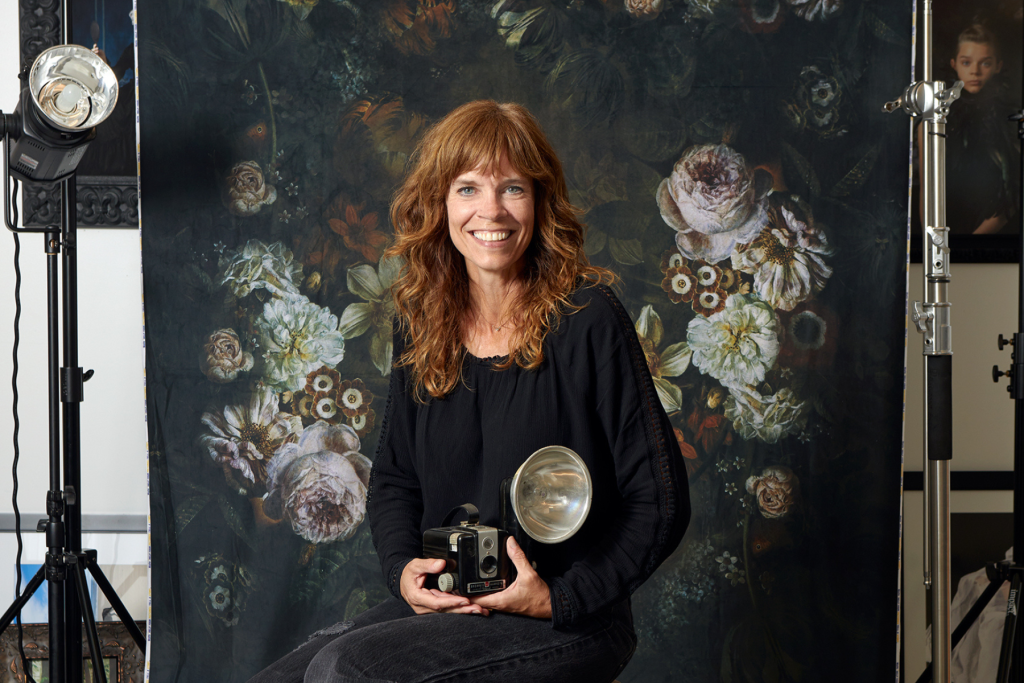 ARTS 14C studio artist Megan Maloy smiling and holding a vintage camera with flash, seated in her studio before a large, dark floral artwork and surrounded by photography equipment.