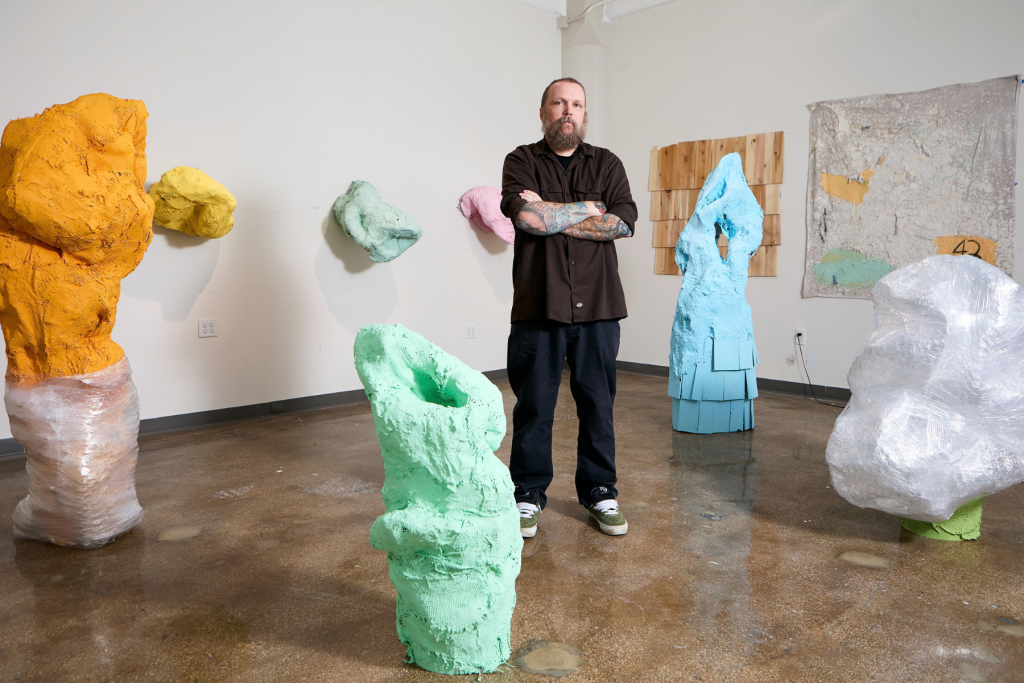 Artist Michael Conlan stands with arms crossed in his ARTS 14C studio, surrounded by colorful, rough-textured abstract sculptures in orange, green, blue, pink, and translucent white.