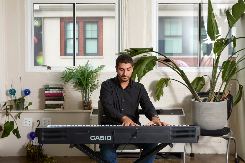 Artist Nahed Elrayes plays an electronic keyboard in a bright ARTS 14C studio, framed by large windows, leafy plants, and a stack of books.