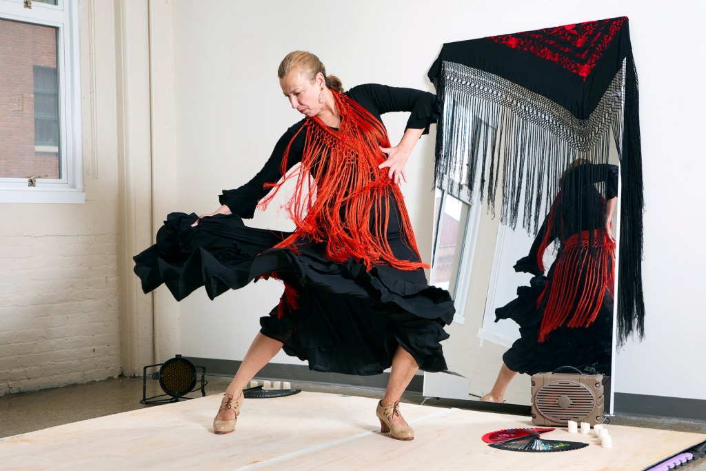 Artist Natallia Basava performs a flamenco pose in a black ruffled dress with a vivid red fringed shawl, reflected in a large mirror draped with a black-and-red fringed textile.