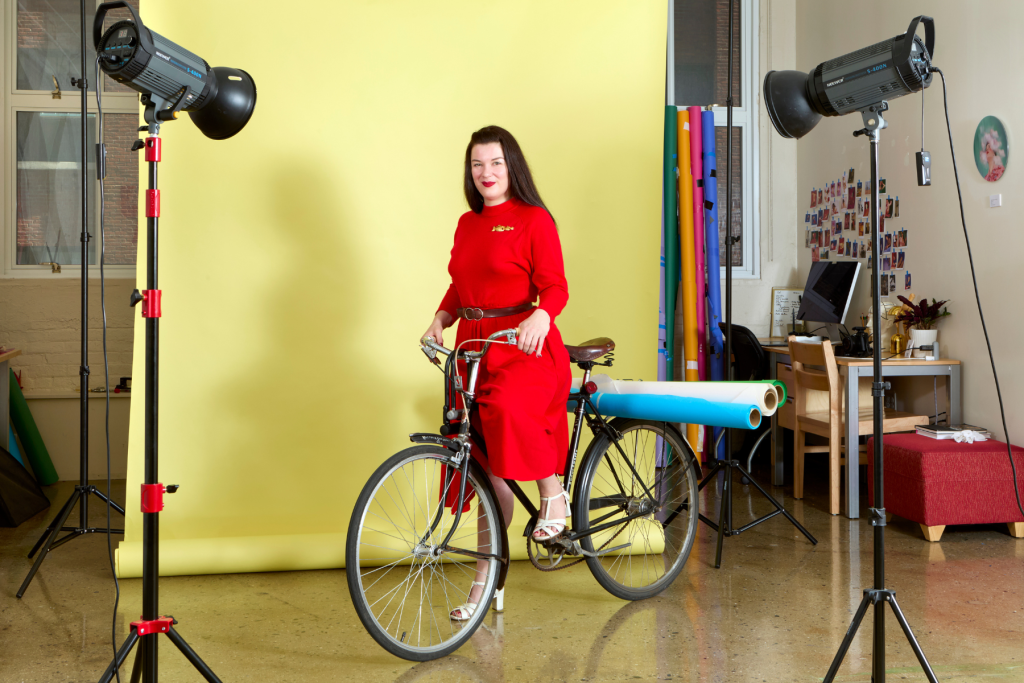 Artist Nathalie De Zan poses with a bicycle in her ARTS 14C studio, wearing a bright red dress against a yellow photography backdrop, surrounded by studio lights and rolls of colorful paper.