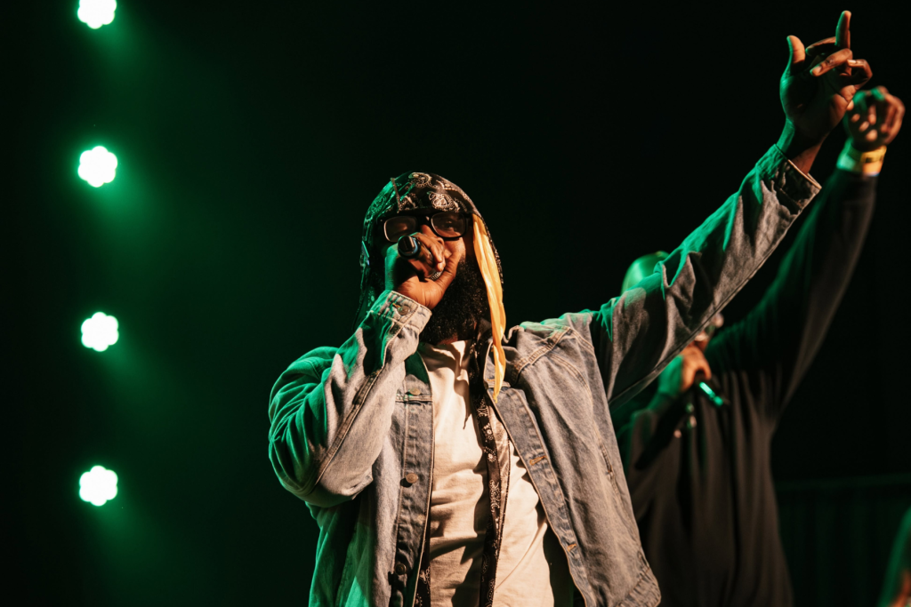ARTS 14C Fellowship Artist Petey performing onstage with a microphone, raising one arm toward the audience under dramatic green stage lighting.