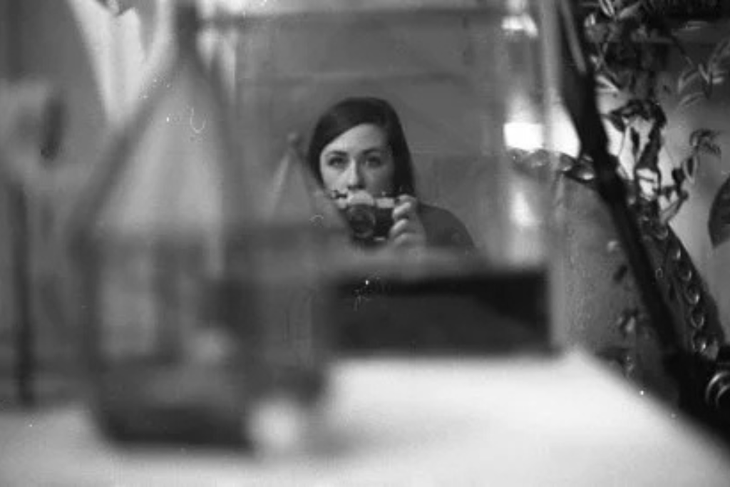 Black-and-white self-portrait of an artist holding a camera and looking into a mirror, partially obscured by soft, out-of-focus reflections and objects in the foreground.