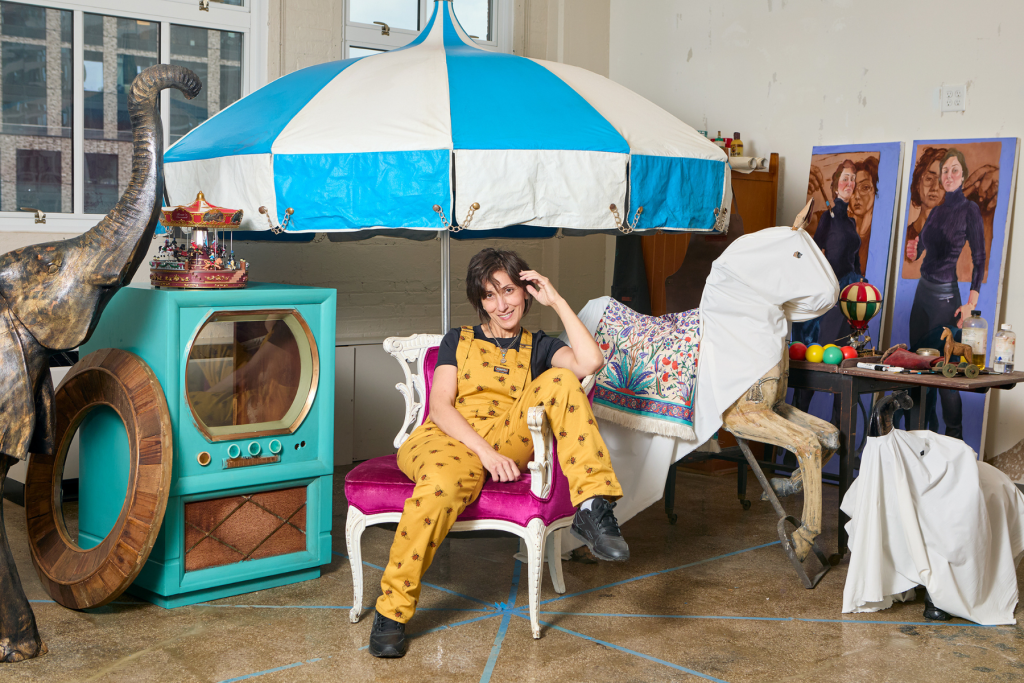 Artist Riham ElSadany sits beneath a large blue-and-white canopy in her ARTS 14C studio, surrounded by whimsical sculptural objects, a carousel horse, colorful props, and figurative paintings.
