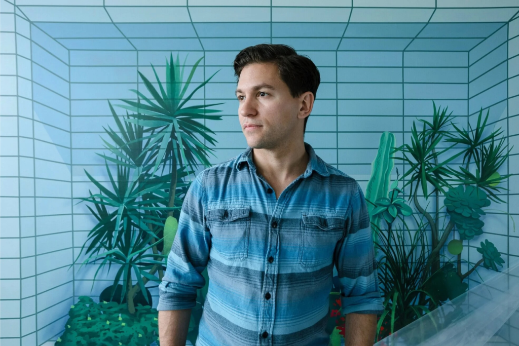ARTS 14C Fellowship Artist Robert Minervini standing before a blue-green geometric mural depicting lush tropical plants within a tiled architectural space.