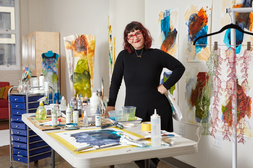 Artist Samar Hussaini stands in her ARTS 14C studio surrounded by colorful abstract works, textile pieces, and a worktable filled with paints, brushes, and other art materials.
