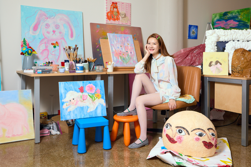 Artist Sherly Fan seated in a colorful studio surrounded by whimsical pastel paintings, art supplies, and sculptural works featuring playful animals and faces.