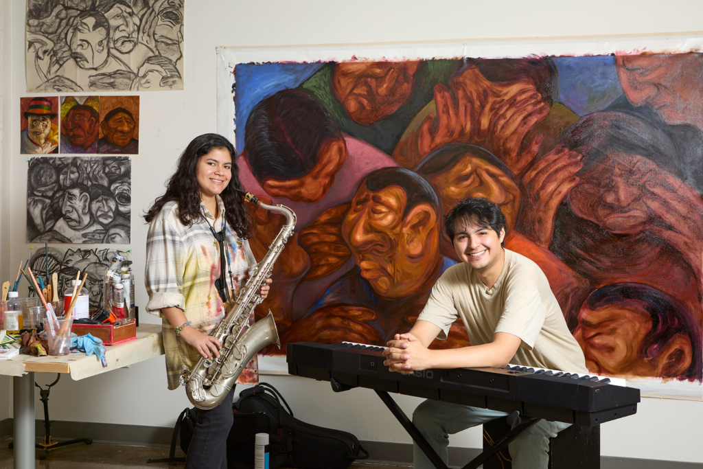 Artists Sofia Carrasco Iglesias and Victor Sotomayor pose with a saxophone and keyboard in their studio, surrounded by expressive paintings of faces and art supplies.