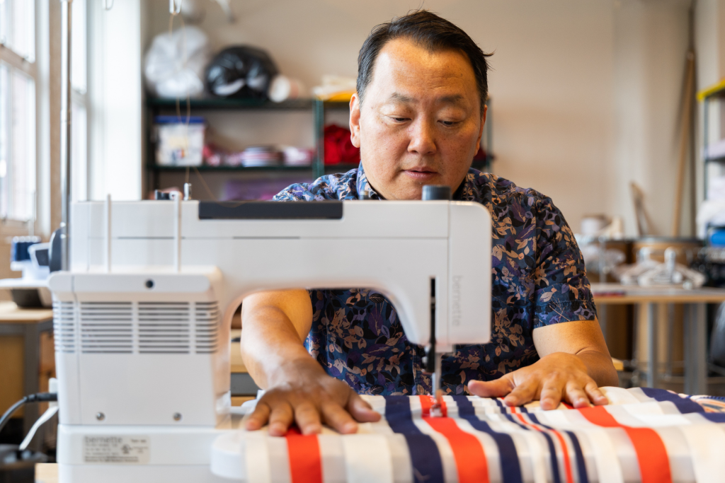 ARTS 14C Fellowship Artist Sok Song works at a sewing machine in his studio, carefully guiding red, white, and blue striped fabric beneath the needle.