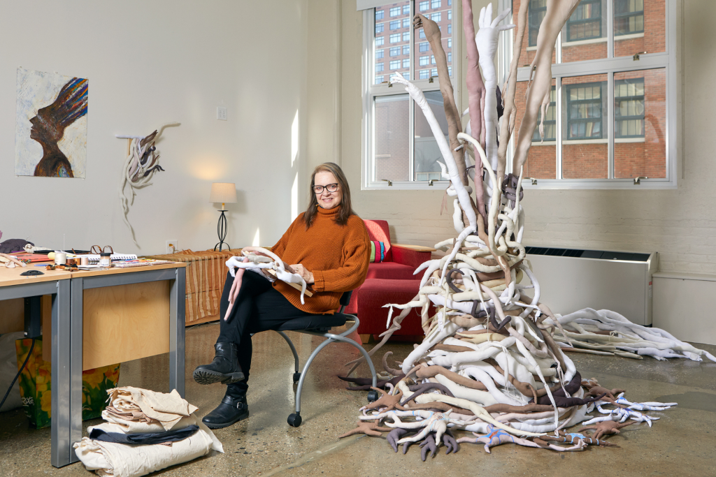 Artist Tracy Leavitt sits in her ARTS 14C studio beside a large, branching textile sculpture composed of intertwined fabric forms in soft neutral tones, with sewing materials and additional works surrounding her.