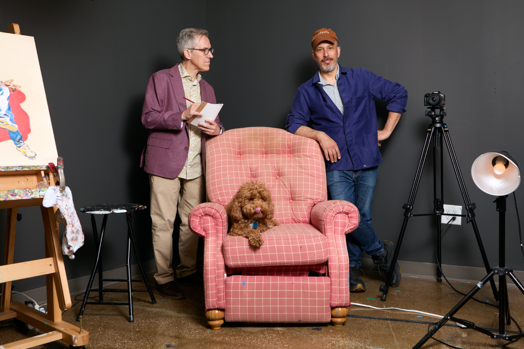 Writer Tris McCall and painter Ben Fine stand in their ARTS 14C studio beside a pink armchair occupied by a small brown dog, surrounded by an easel, camera, and studio lighting against a dark wall.