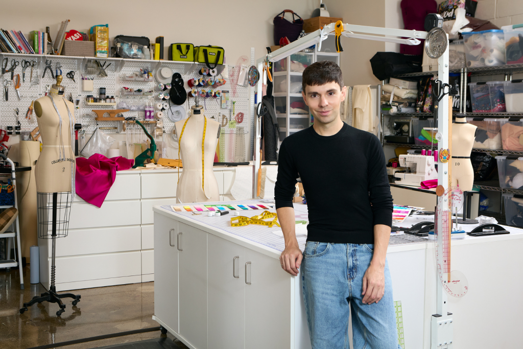 ARTS 14C Studio Artist Tyler Neasloney standing in his fashion design studio, surrounded by dress forms, sewing equipment, colorful fabrics, thread, patterns, and tailoring tools.