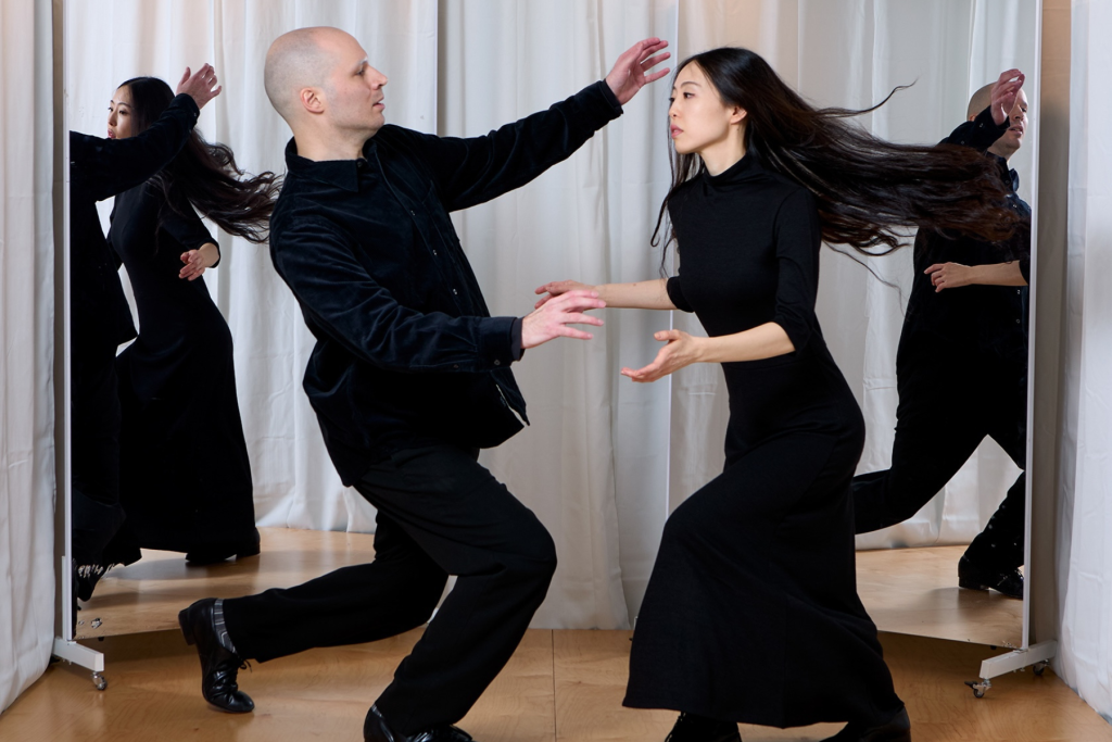 Dancers Warren Craft and Rena Kinoshita perform a dynamic movement together in black clothing, their gestures and flowing motion reflected in large mirrors behind them.