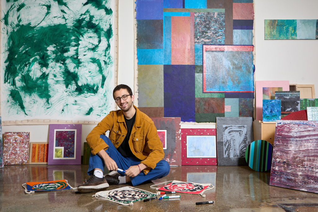 Artist William Masters sits cross-legged on the floor of his ARTS 14C live/work studio, surrounded by colorful abstract paintings and works in progress, with large layered compositions filling the wall behind him.