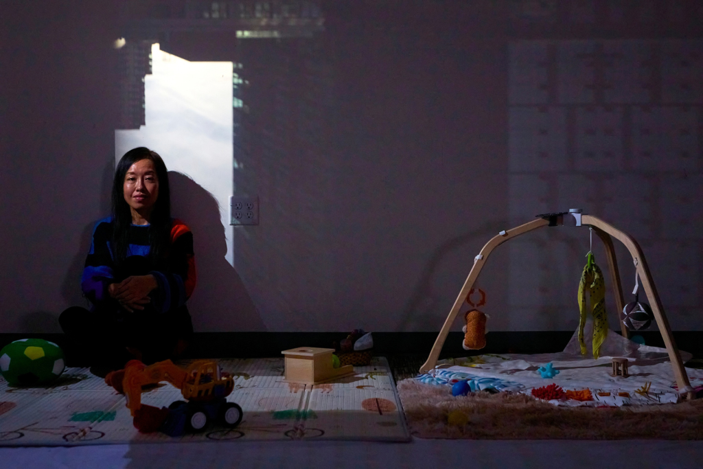 Artist Yu-Chen Chiu sits in a darkened installation space surrounded by children’s toys and sensory objects, with a projected cityscape casting light and shadow across the wall behind her.