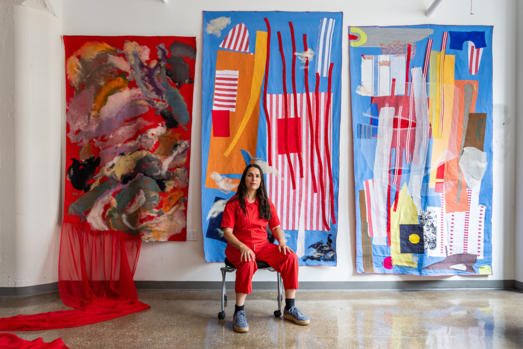 Artist gwen charles sits in her studio wearing red, surrounded by three large textile works featuring bold abstract forms, layered fabrics, and vivid red, blue, yellow, and orange.