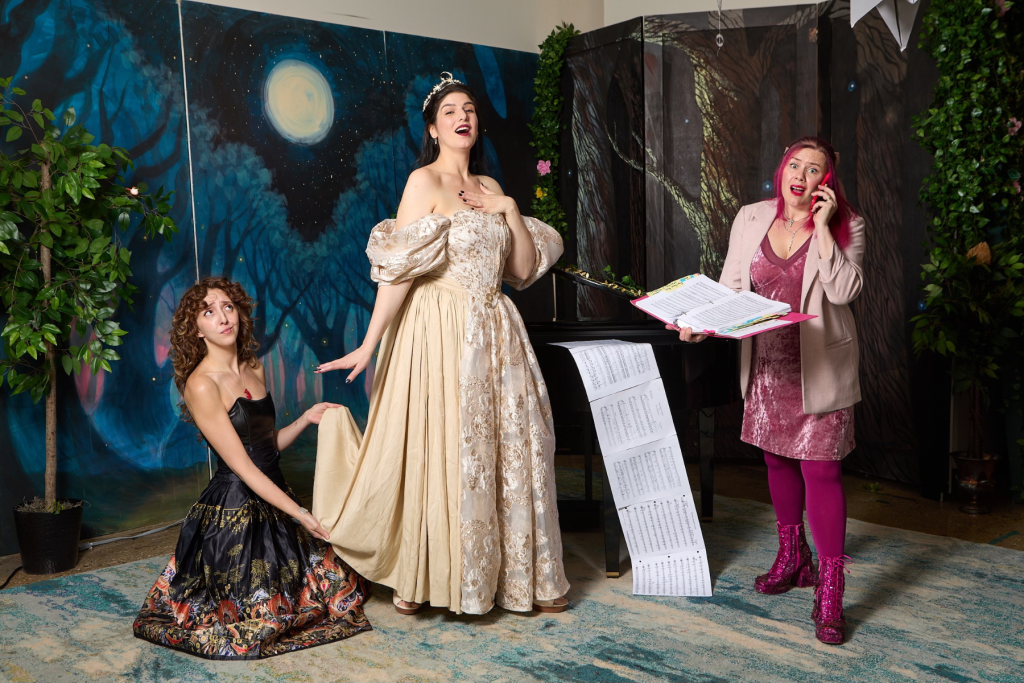The three founders of Undead Arts pose theatrically in a woodland-inspired set: one kneels in a dark floral gown, another stands in an ornate cream gown and tiara, and a third holds a script while talking on a pink phone beside a piano and cascading sheet music.
